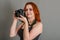 Smiling woman with red hair holds a modern camera in her hands. Gray background, creativity, photographer, beautiful