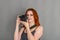 Smiling woman with red hair holds a modern camera in her hands. Gray background, creativity, photographer, beautiful