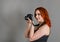 Smiling woman with red hair holds a modern camera in her hands. Gray background, creativity, photographer, beautiful