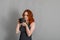 Smiling woman with red hair holds a modern camera in her hands. Gray background, creativity, photographer, beautiful