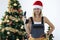 Smiling woman repairman in uniform and santa claus hat holding drill in hands near christmas tree