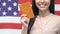 Smiling woman showing passport against American flag, getting USA citizenship