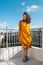 A smiling woman stands on a roof overlooking the city and the wind inflates her dress