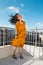 A smiling woman stands on a roof overlooking the city and the wind inflates her dress