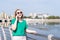 A smiling woman in sunglasses, talking on the phone with friends gesturing, making date on the riverbank in the city.