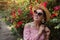 Smiling woman wearing straw hat walking by blooming roses in summer garden. Girl enjoys flowers in park. Summer outfit.