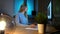 Smiling woman working on computer at night. Smiling female in checkered shirt sitting at lit by small lamp wooden desk