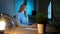 Smiling woman working on computer at night. Smiling female in checkered shirt sitting at lit by small lamp wooden desk