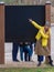 Smiling woman in yellow jacket points fingers at empty black stand in autumn city park.