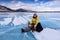 Smiling woman in a yellow jacket sits on the blue ice of lake Baikal and changes her red boots to ice skates