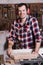 Smiling woodworker. Cheerful young male carpenter leaning at the circular table with wooden plank