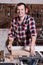 Smiling woodworker. Cheerful young male carpenter leaning at the circular table with wooden plank