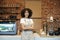 Smiling young african american lady in apron stands near bar counter with coffee making equipment and looks at camera