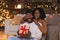 Smiling young african american wife closes eyes to husband, shows silence sign and gives gift box