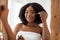 Smiling young black lady in towel using facial serum near mirror, taking care about her beauty at home