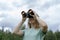 Smiling Young blonde woman bird watcher in cap and blue t-shirt looking through binoculars at cloudy sky in summer forest