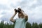 Smiling Young blonde woman bird watcher in cap and blue t-shirt looking through binoculars at cloudy sky in summer forest