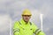 Smiling young engineer with hard hat and protective clothing in front of blurred wind turbines