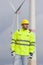 Smiling young engineer with hard hat and protective clothing in front of blurred wind turbines