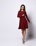 Smiling young female in a rich red long-sleeved flying gown and nude shoes standing cross-legged putting hand on hip,