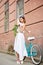 Smiling young female in white dress posing with peonies near blue bike in front of red historical building
