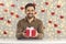 Smiling young man holding Valentine present sitting at table against hearts and lights wall decor