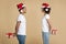Smiling young man and woman in Santa Claus hats look at each other and hold surprise gifts behind backs