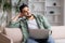 Smiling young muslim man with beard watch at free space, sits on sofa with laptop in living room interior