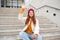 Smiling young redhead girl waits someone on stairs outdoors, waves hand at friend, holds smartphone, says hello