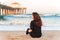 A smiling young woman with long hair sits on the sand in front of a pier on Manhattan Beach in Los Angeles in the early morning