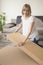 Smiling young woman opens cardboard boxes in the living room. Sofa and flower in the background.