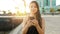 Smiling young woman sending message with smart phone at sunset with seaside town on the background. Positive trendy girl blogging