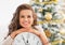 Smiling young woman showing clock in front of christmas tree