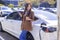 Smiling young  woman standing on city parking near electric car, charging automobile battery from small city station, drinking cof