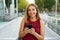 Smiling young woman is walking in the street with phone. She is holding gadget in hand and looking at camera