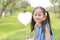 Smilling little Asian girl holding blank heart label lying on green grass at summer garden