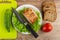 Smoked pork neck with lettuce and knife in plate, cucumber on cutting board, pieces of bread, tomato on table. Top view