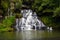 Smooth milky water by low shutter. Beautiful and large milky waterfall surrounded by trees, grass, dark stone, lake