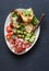 Snack bowl - prosciutto, olives, cherry tomatoes, avocado dip, grilled mozzarella spinach sandwiches on dark background, top view.