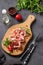 Snack wooden board with bacon or prosciutto on a dark background with spices, tomatoes and herbs