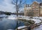 Snow blankets the ground while a bright blue sky reflects in the mirror-like surface of a small pond.