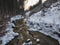 Snow covered forest water stream creek with trees, branches and stones, idyllic winter landscape in golden hour sun