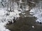 Snow covered forest water stream creek with trees, branches and stones, idyllic winter landscape in golden hour sun