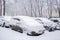 Snow-covered parked cars standing in snowdrift during snowfall.