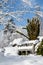 Snow covered wood bench in a snowy winter garden landscape, trees and bushes against a blue sky and white clouds