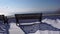 Snow is falling on empty city park. A lonely snow-covered bench stands near sea. Christmas winter new year scenery