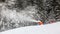Snow gun spraying artificial ice crystals to ski piste, snowmaking in winter sports resort, trees in background