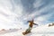 A snowboarder in a ski mask and a backpack is riding on a snow-covered slope leaving behind a snow powder against the