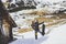 Snowboarder wearing black jacket, mask with snowboard close authentic wooden house.Man looking at panoramic mountans