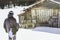 Snowboarder wearing black jacket, mask with snowboard close authentic wooden house.Man walking with snow board desk